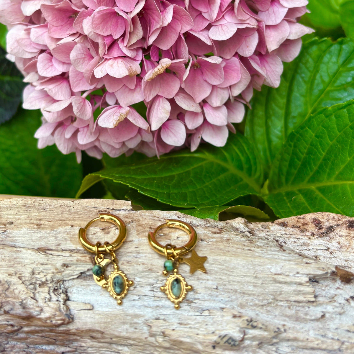 A pair of gold hoop earrings with dangling charms, including a star and a turquoise-blue oval stone, displayed on a piece of weathered wood with green leaves and pink flowers in the background.