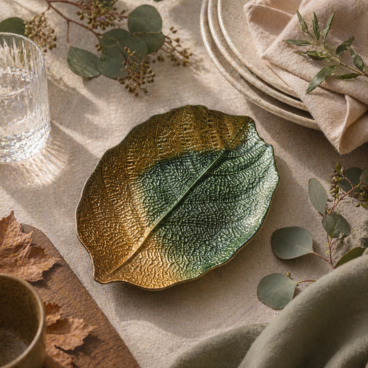 Decorative leaf-shaped dish on a table with greenery and a glass.