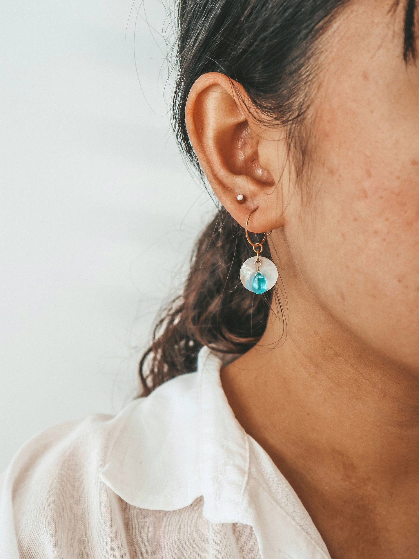 Close-up of an ear wearing a hoop earring with a turquoise gemstone on a plain background