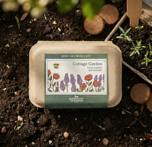 A mini cottage garden starter kit by Farmer's Essentials, packaged in a brown paper tray, placed on dark soil with small seedlings and wooden potting sticks.