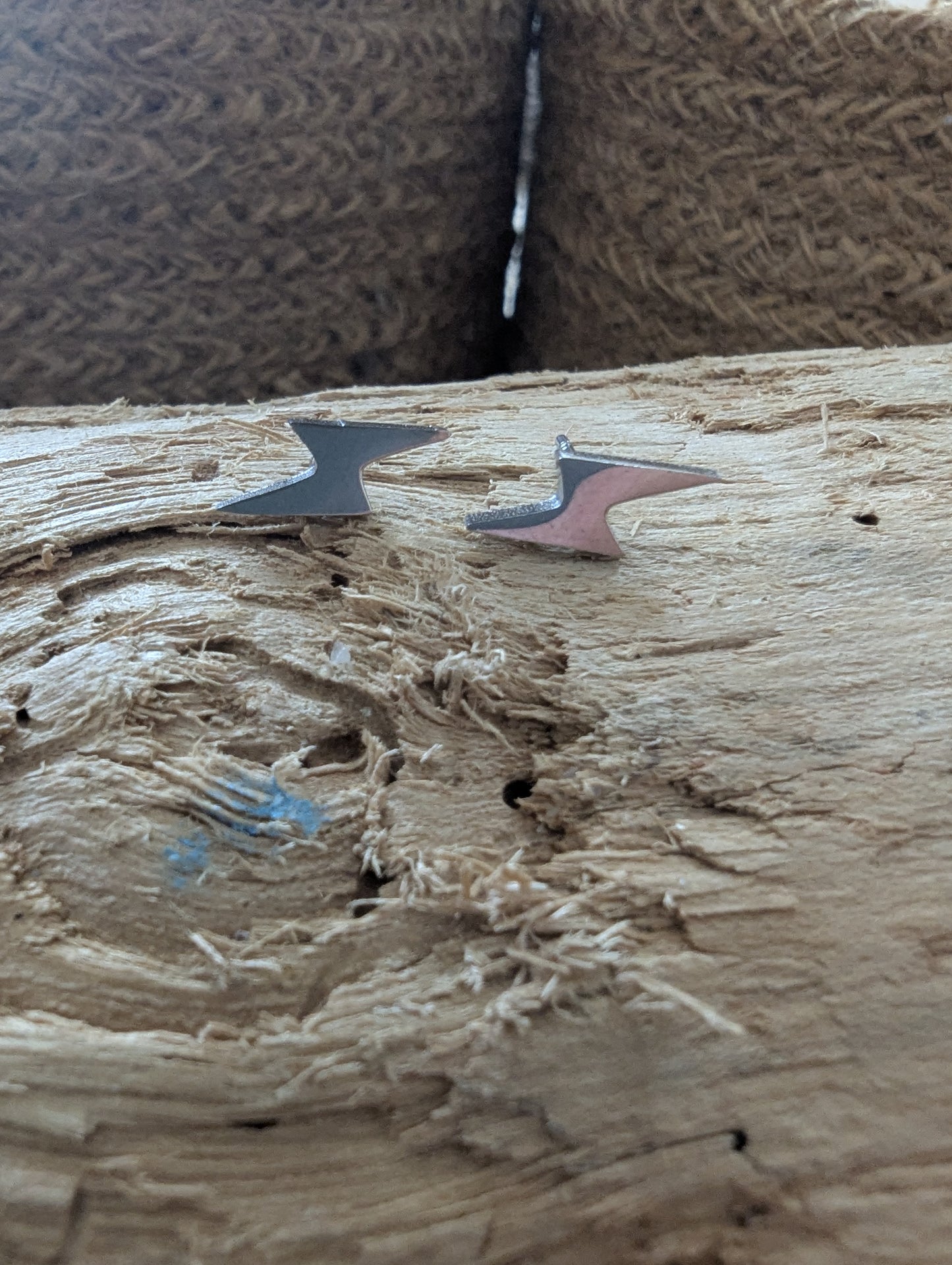 Two small abstract metal arrowhead-shaped earrings or charms resting on a rough wooden surface, with a textured brown woven background behind them.