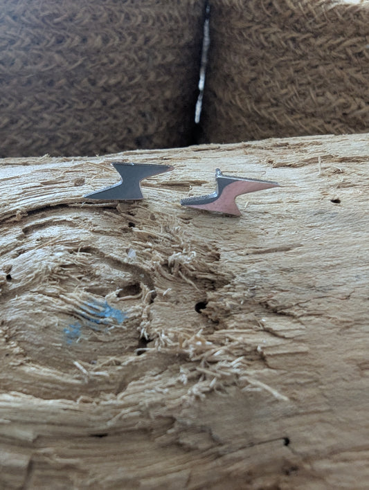 Two small abstract metal arrowhead-shaped earrings or charms resting on a rough wooden surface, with a textured brown woven background behind them.