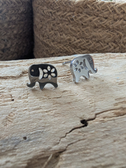 Two small elephant-shaped stud earrings displayed on a rustic wooden surface, with a woven brown background. One earring is dark silver/black with white paw-print cutouts, and the other is silver with dark paw-print cutouts.