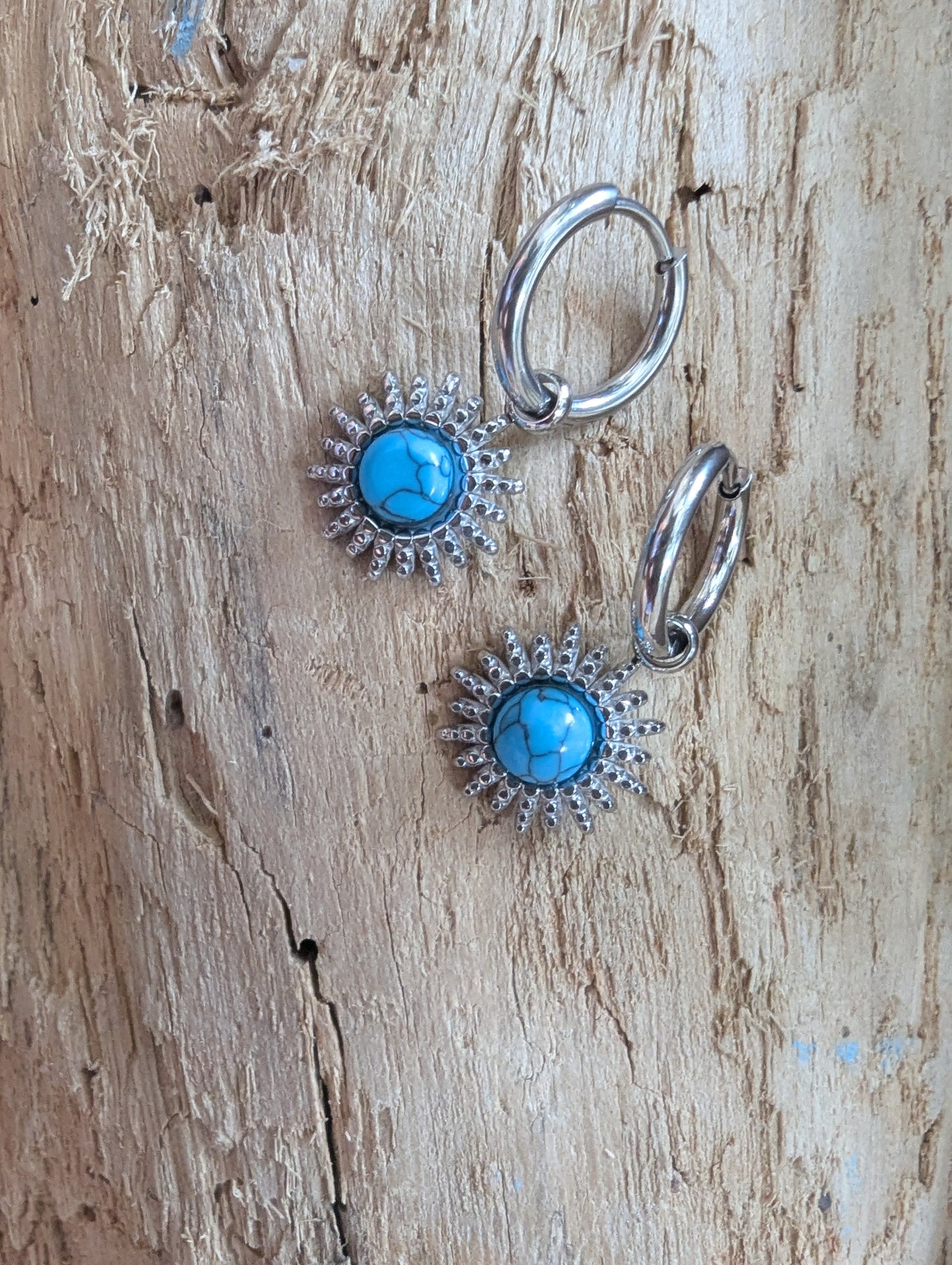 A pair of silver hoop earrings with round turquoise blue center stones surrounded by a sunburst halo of small clear rhinestones, displayed on a textured light brown wood background.