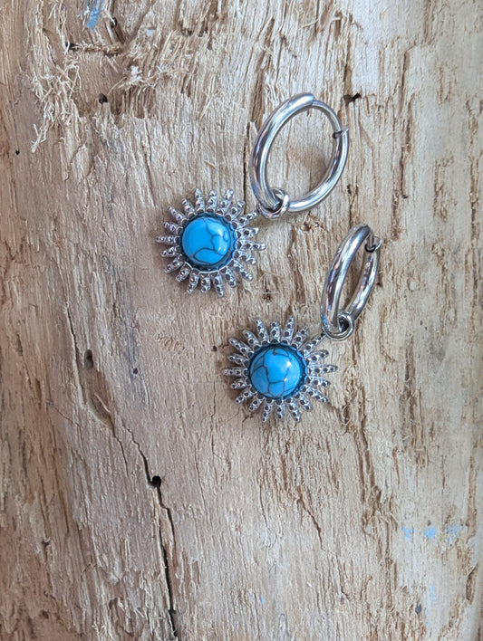 A pair of silver hoop earrings with round turquoise blue center stones surrounded by a sunburst halo of small clear rhinestones, displayed on a textured light brown wood background.