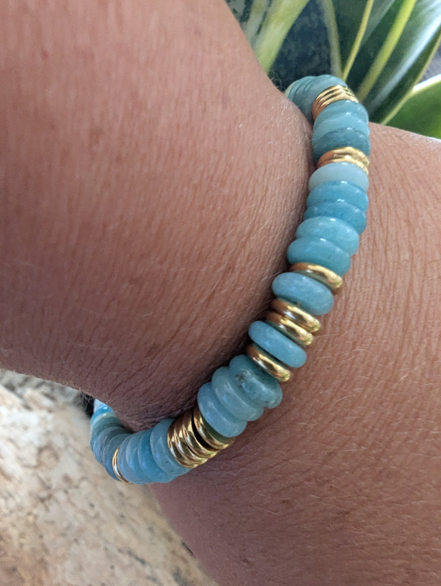 A close-up of a beaded stretch bracelet worn on a wrist. The bracelet features light blue stone beads separated by shiny gold metallic spacers.