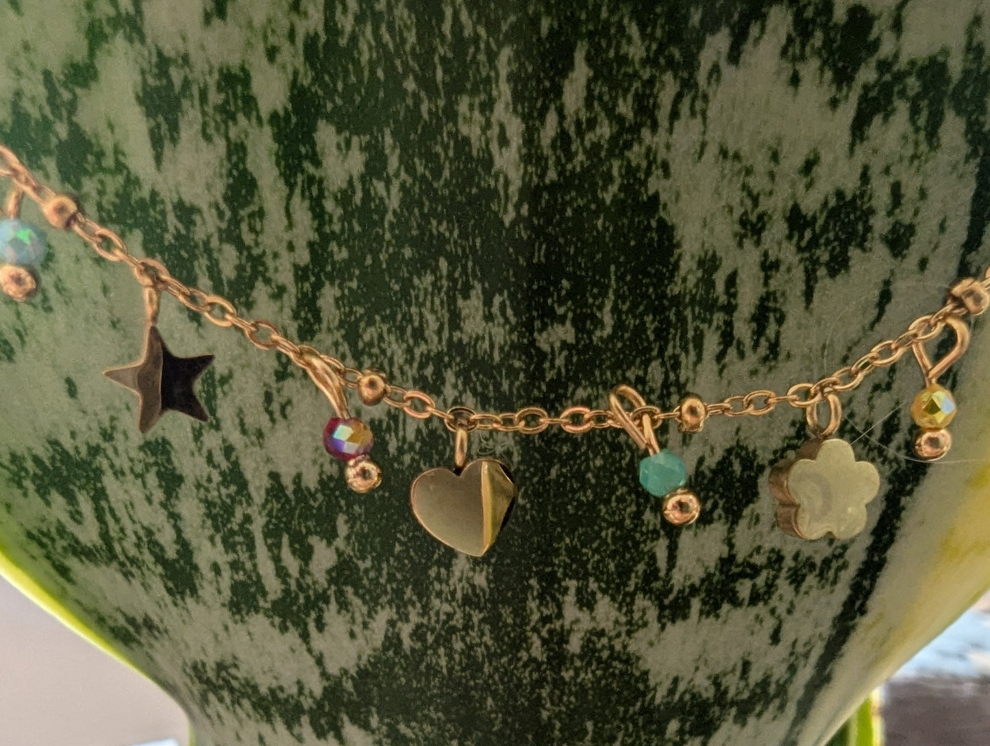 A gold-tone charm necklace draped across a green and white variegated snake plant pot. The necklace features multiple charms including a star, a heart, a clover, and small colorful beads.