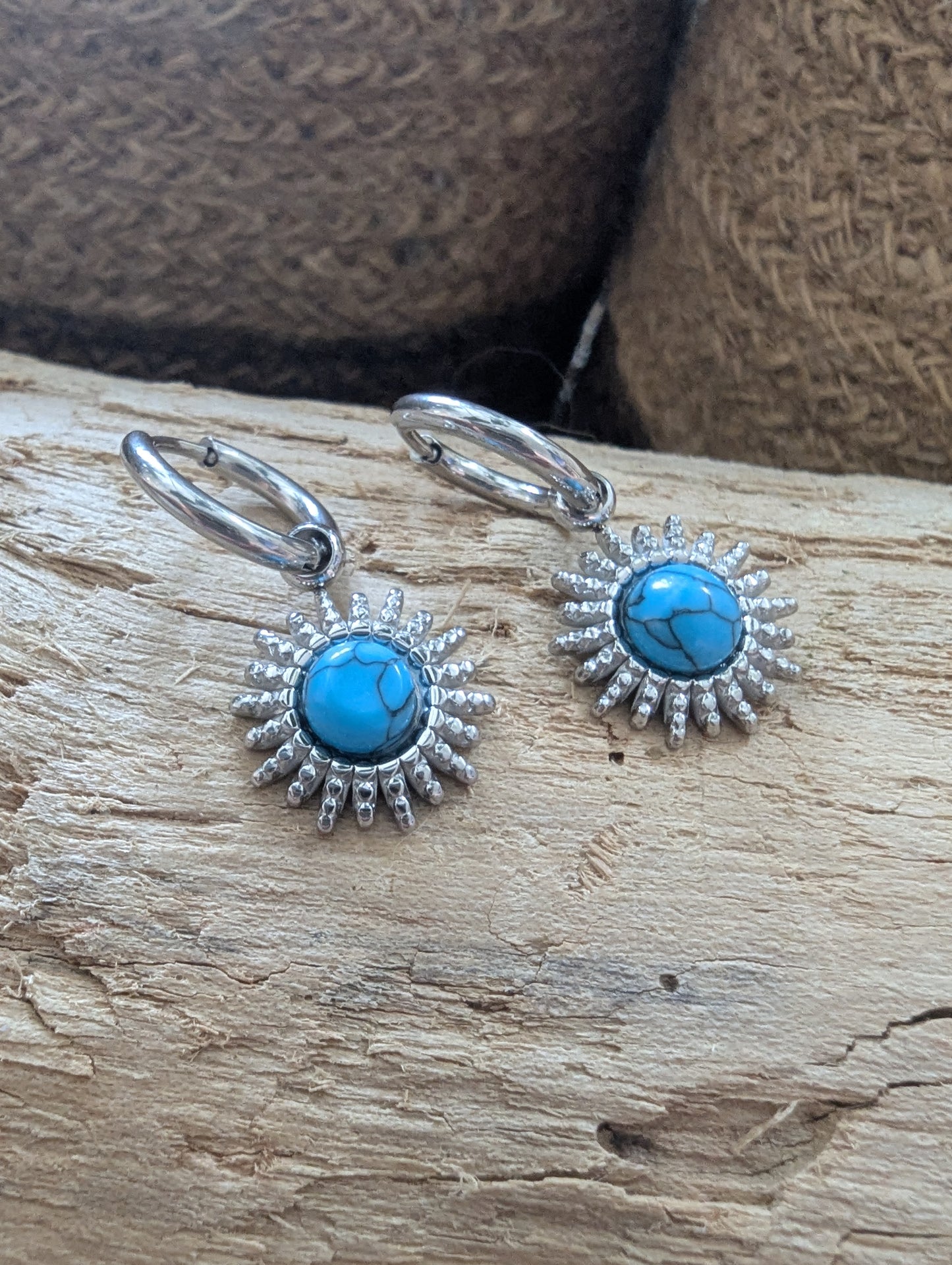 A pair of sterling silver drop earrings with round turquoise cabochons and sunburst detailing, displayed on a piece of natural wood with a woven brown background.