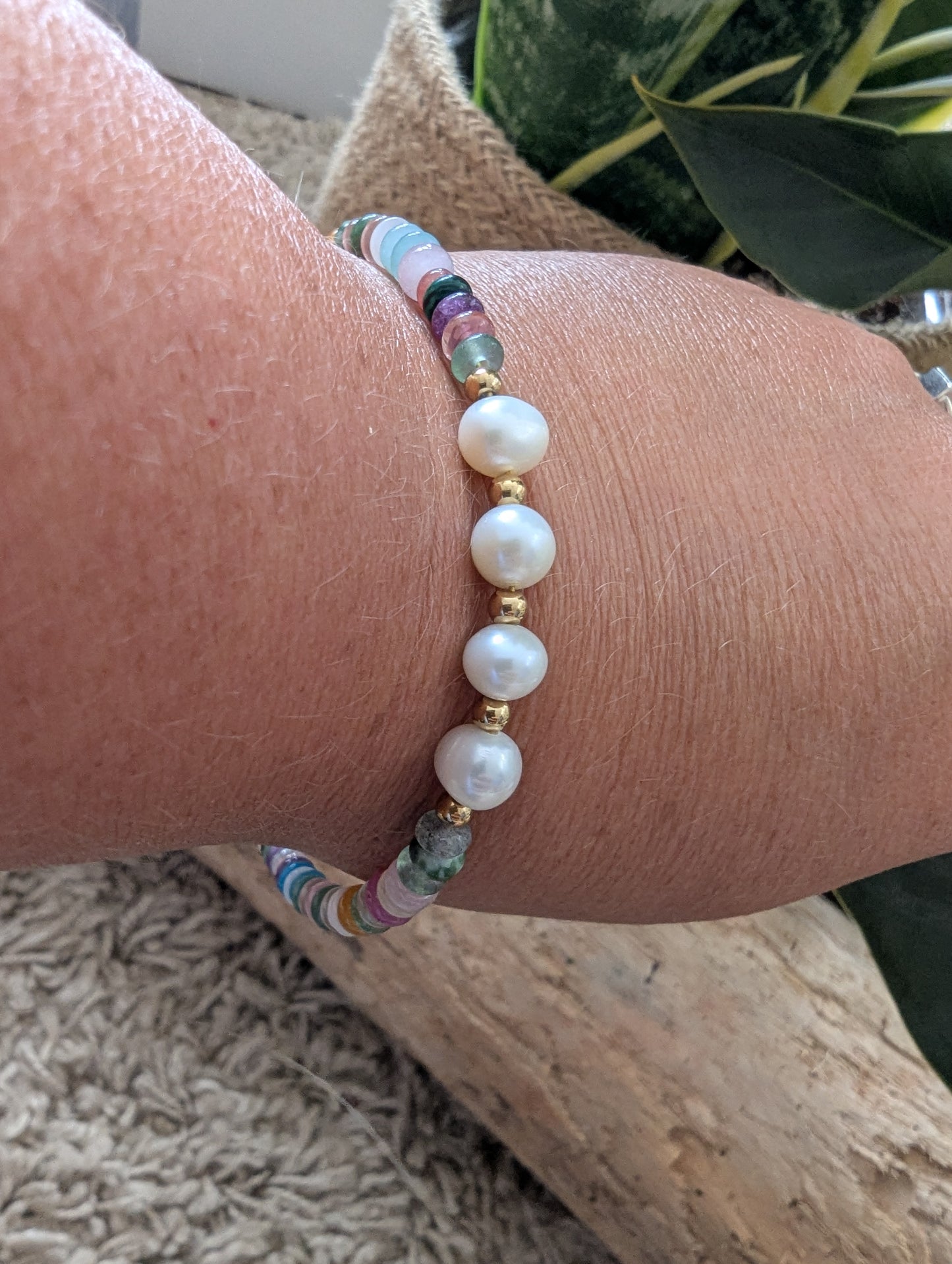 A close-up of a person's wrist wearing a beaded bracelet with white pearl-like beads, gold spacer beads, and small multicolored faceted beads, shown in a natural setting with wood and greenery in the background.