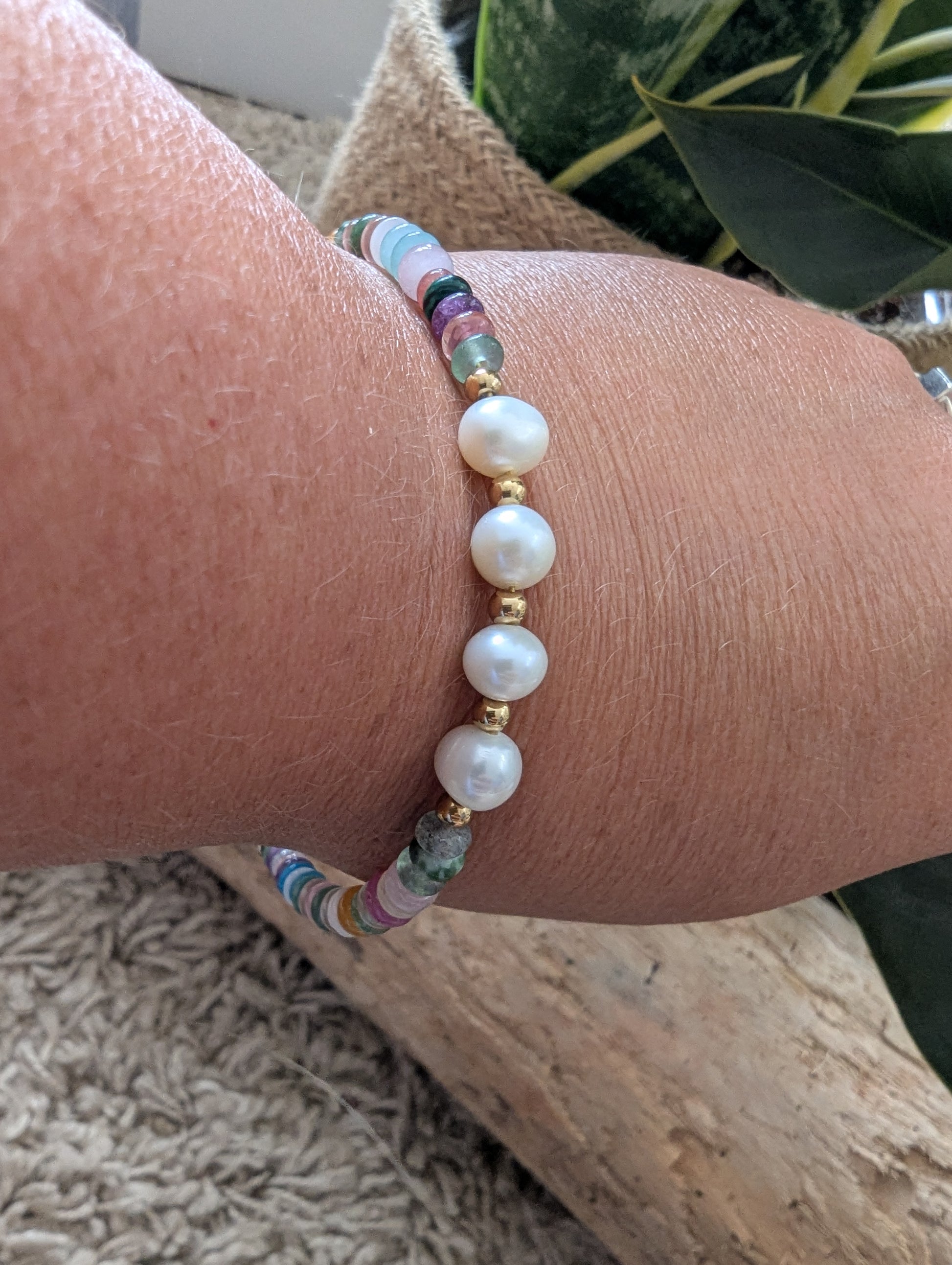 A close-up of a person's wrist wearing a beaded bracelet with white pearl-like beads, gold spacer beads, and small multicolored faceted beads, shown in a natural setting with wood and greenery in the background.