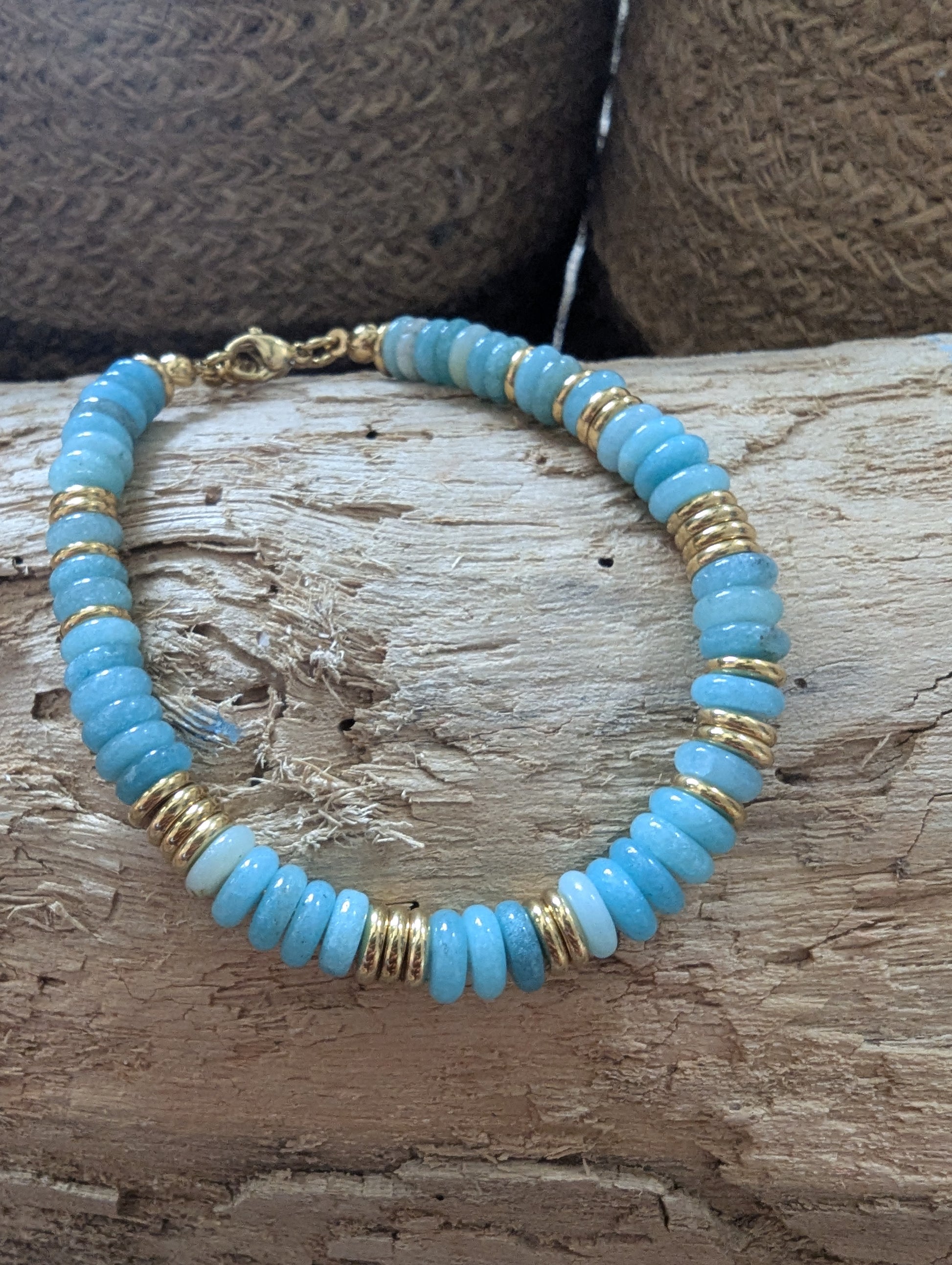 A beaded bracelet featuring light blue stone beads, white beads, and textured gold spacer rings, displayed on a rustic wooden surface.
