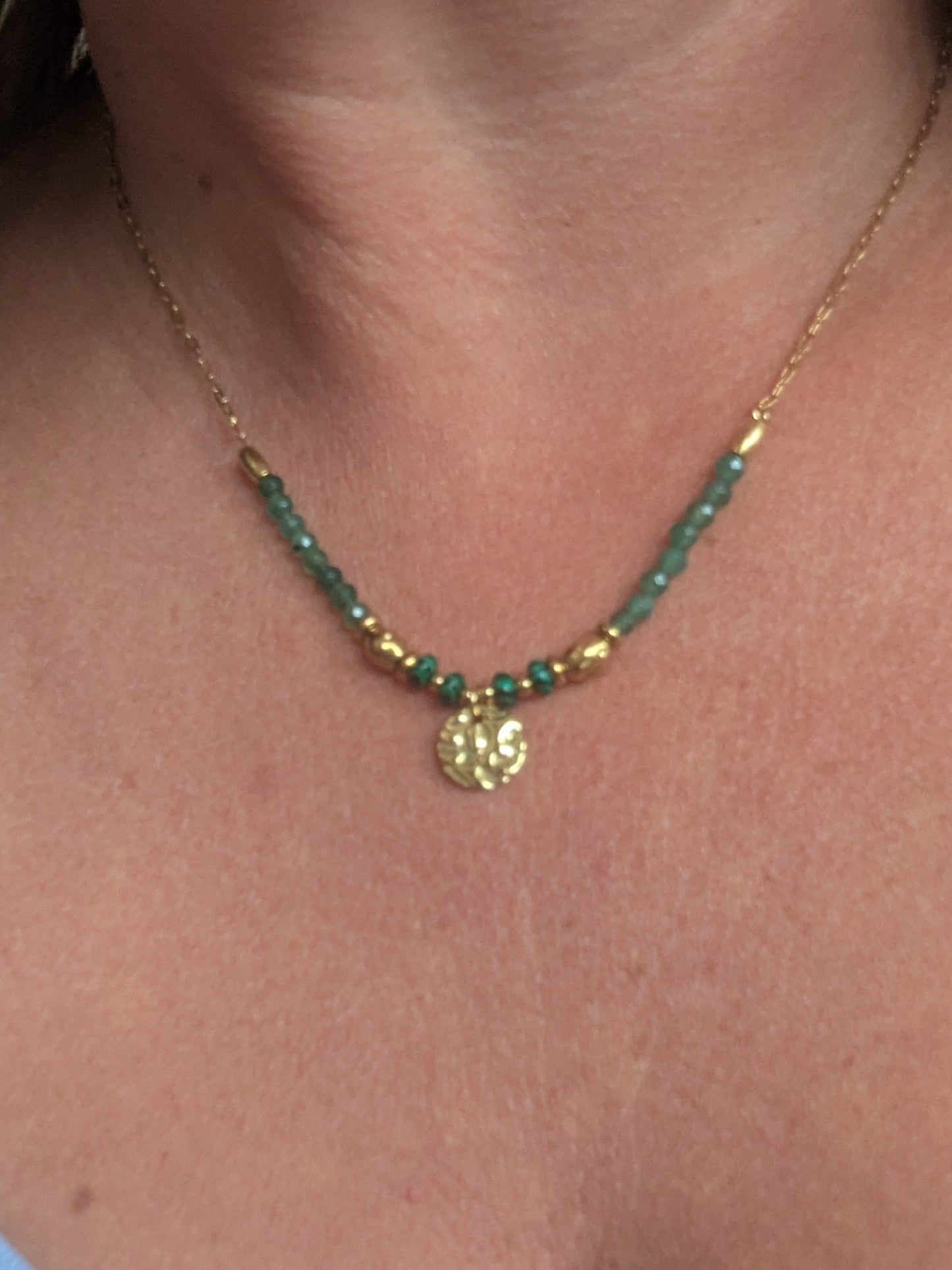 A close-up of a woman's neck and chest wearing a delicate gold chain necklace with green beads and a small gold pendant.