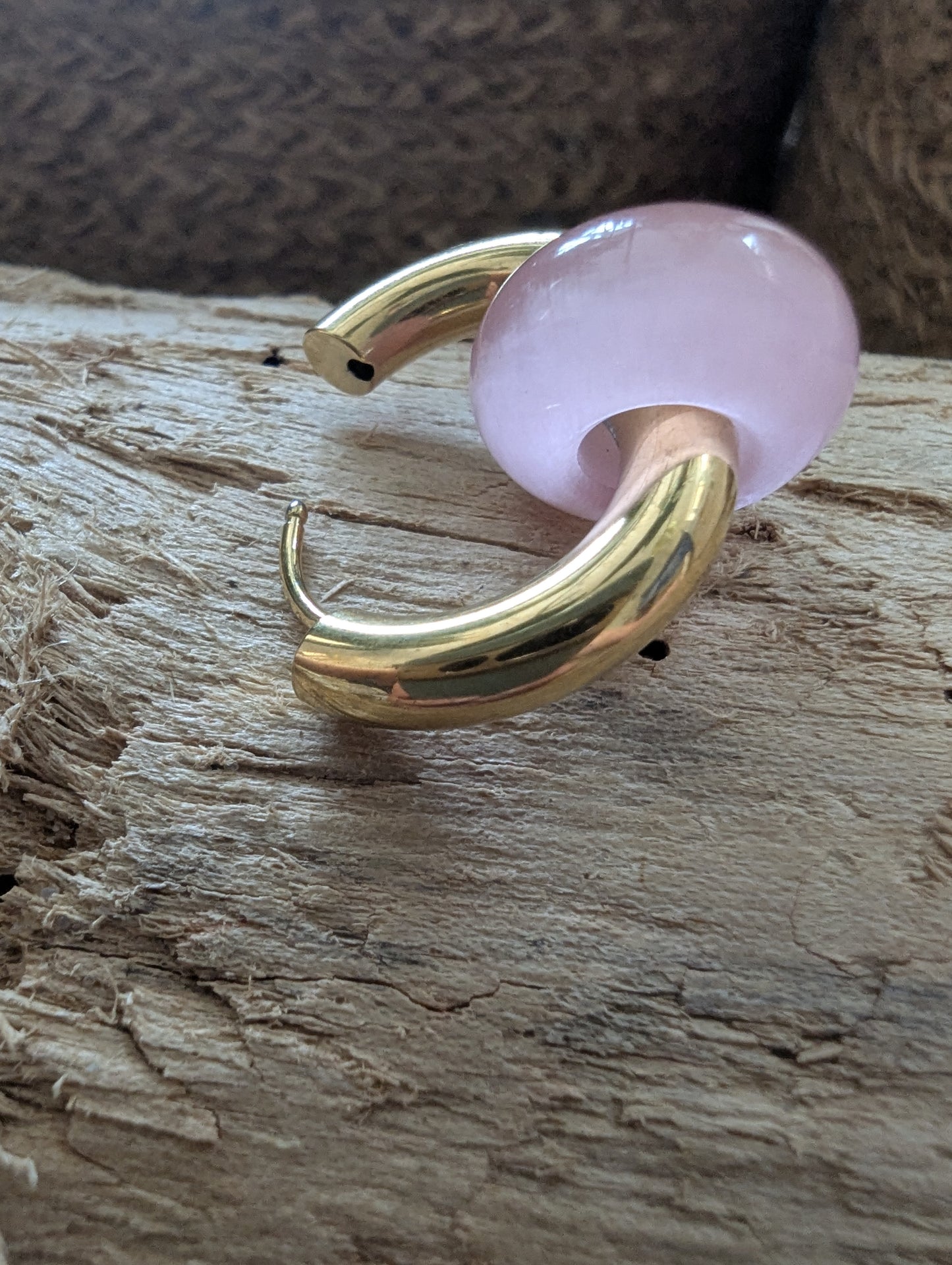 A gold-toned curved hoop earring with a smooth, translucent light pink stone, resting on a textured piece of wood.